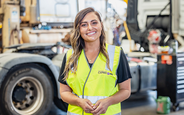 A woman wearing a yellow vest stands in front of a truck, representing the Capricorn team with pride and determination.