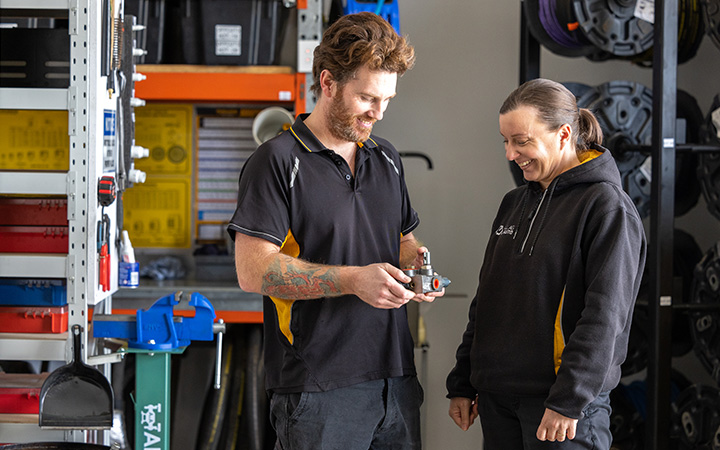 Two mechanics in a storage room, one of them holds a vehicle part.