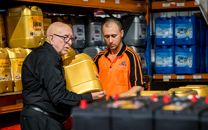 Two people in a warehouse discussing and handling large yellow containers of motor oil, surrounded by shelves stocked with various automotive fluids, representing Capricorn's relationships with Preferred Suppliers.