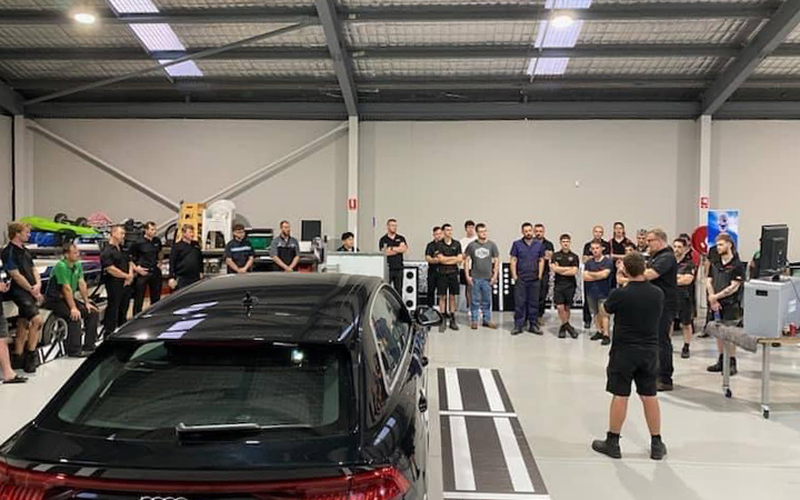 A group of attendees at the Perth Automotive Training Event gathered around a car in a garage setting.