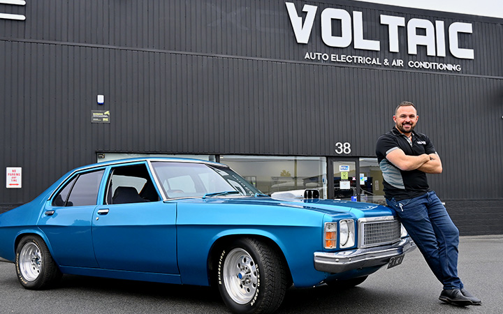 Capricorn Member, Joshua posing and smiling in front his classic blue Holden Kingswood vehicle in front of his workshop
