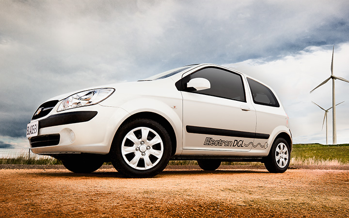 White Blade Electron compact EV with “Electron DGL” branding parked on a rural dirt road, framed by two wind turbines in a grassy field under a partly cloudy sky.