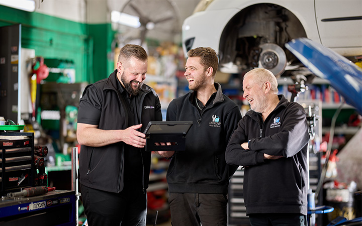 A Capricorn Area Manager and two automotive professionals in a workshop talking and smiling, with a car on a hydraulic lift in the background
