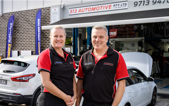 Two automotive technicians, Capricorn Members, in black and red uniforms standing outside a workshop, with two cars and branded signage in the background.