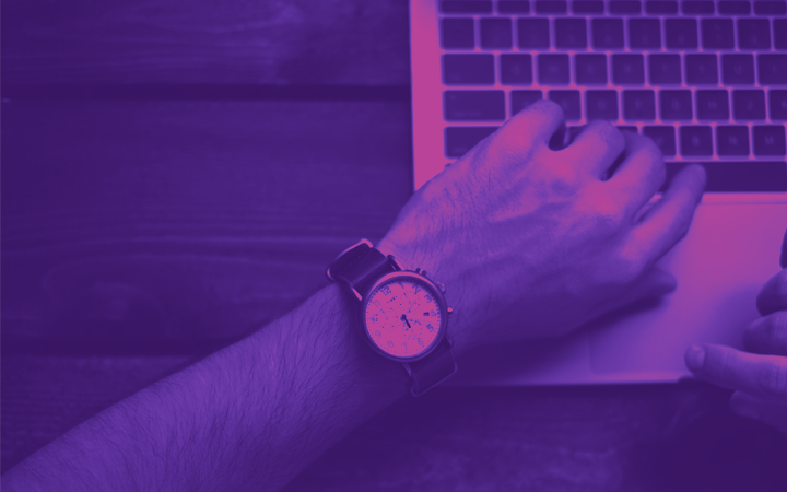 A hand typing on a computer and a watch on the wrist showing 5pm
