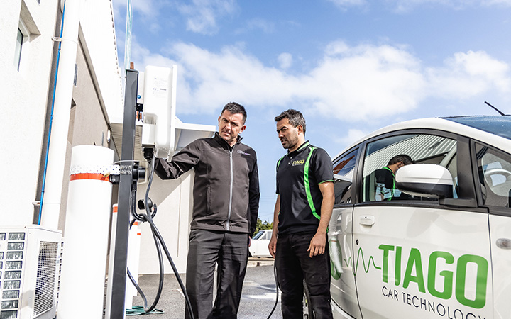 Two men standing next to a white car at a charging station.