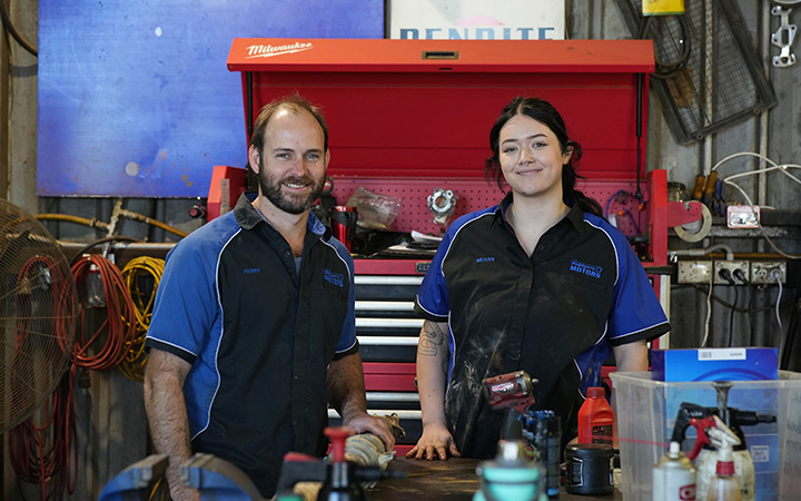 Two automotive technicians in uniforms standing in a workshop, smiling with tools and equipment in the background.