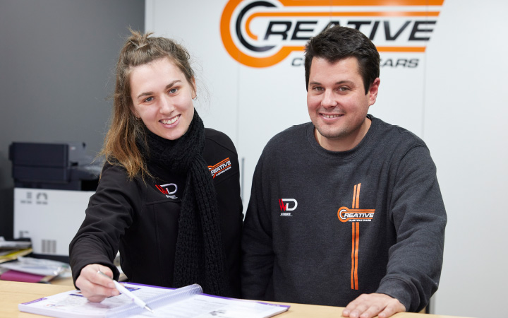Two team members from a Capricorn Member, Creative Custom Cars, reviewing a document at a front counter, with the company logo visible in the background and office equipment behind them.