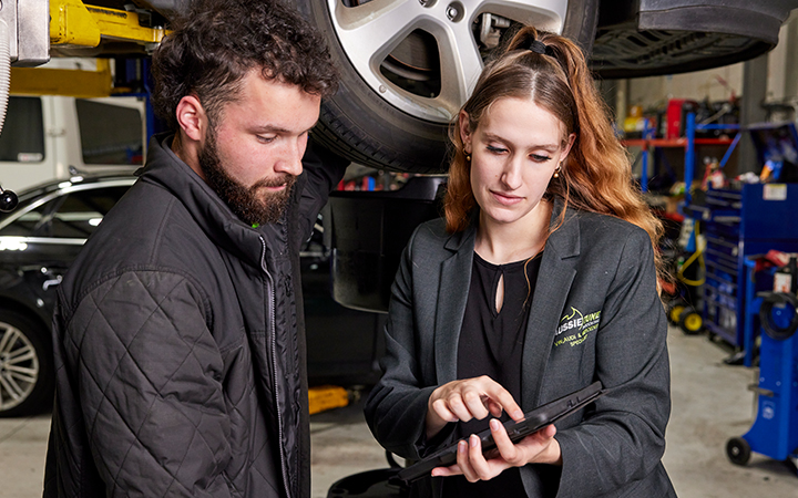 A man and woman in a garage, inspecting a car while examining something on a tablet.