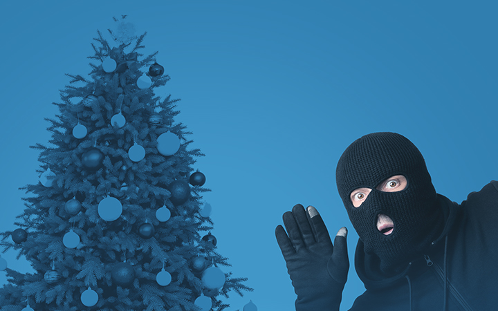 A person in a black mask and hoodie stands by a Christmas tree.