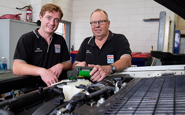 A young apprentice working with an experienced mechanic in a car engine and smiling to the camera