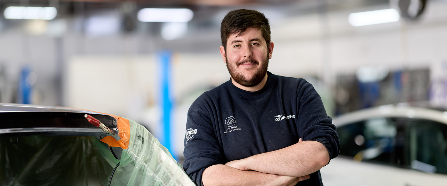 Isaac wearing a branded workshop uniform with arms crossed, positioned beside a car undergoing panel repair.