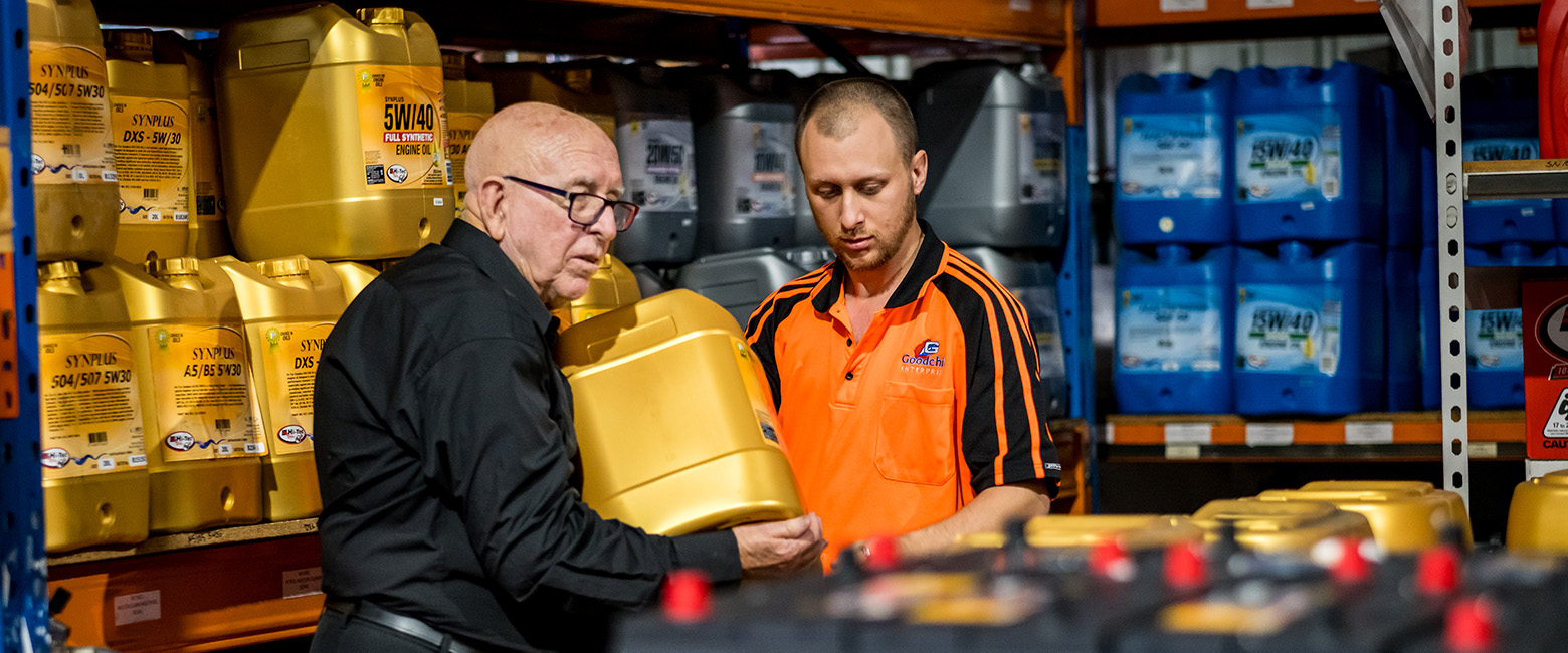 Two people in a warehouse discussing and handling large yellow containers of motor oil, surrounded by shelves stocked with various automotive fluids, representing Capricorn's relationships with Preferred Suppliers.