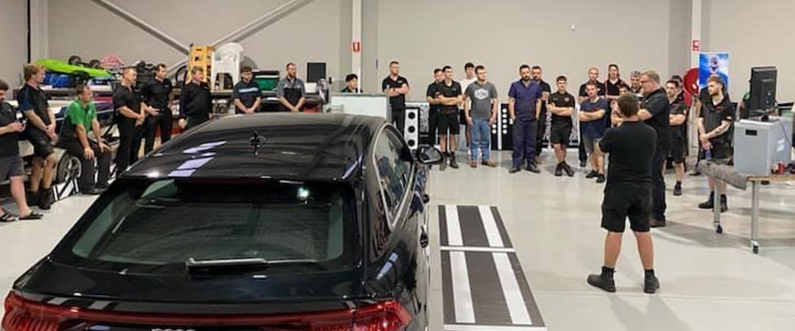 A group of attendees at the Perth Automotive Training Event gathered around a car in a garage setting.
