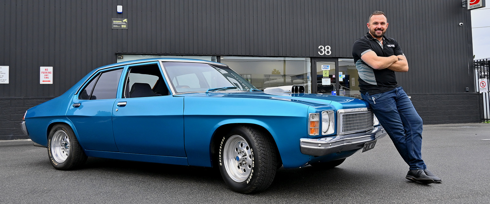 Capricorn Member, Joshua posing and smiling in front his classic blue Holden Kingswood vehicle in front of his workshop