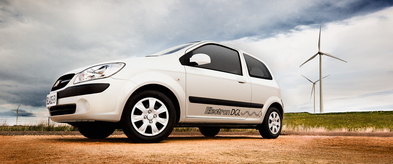 White Blade Electron compact EV with “Electron DGL” branding parked on a rural dirt road, framed by two wind turbines in a grassy field under a partly cloudy sky.