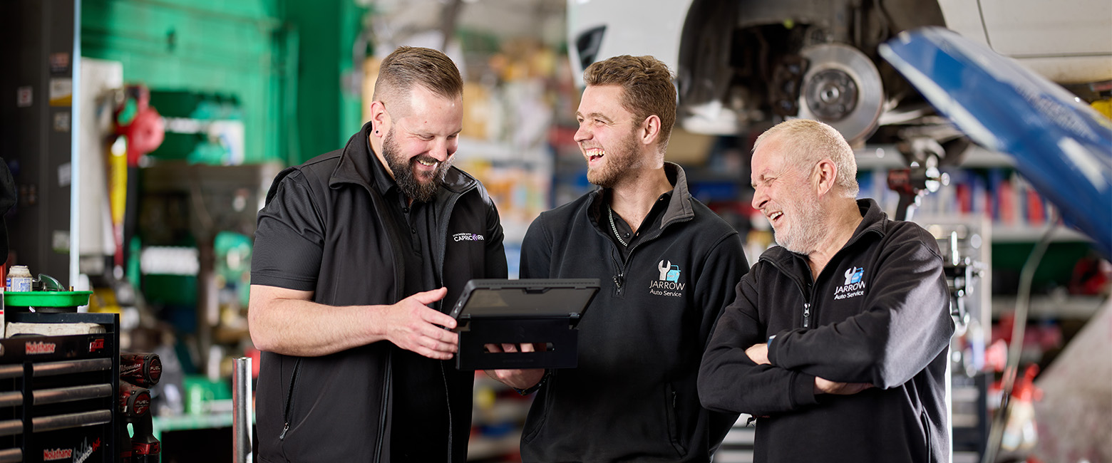 A Capricorn Area Manager and two automotive professionals in a workshop talking and smiling, with a car on a hydraulic lift in the background