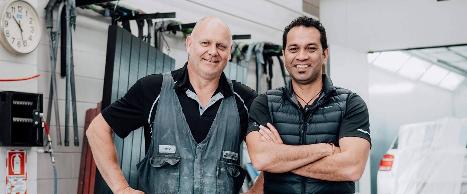 Two auto technicians standing side by side in garage/workshop, both smiling and looking towards the camera