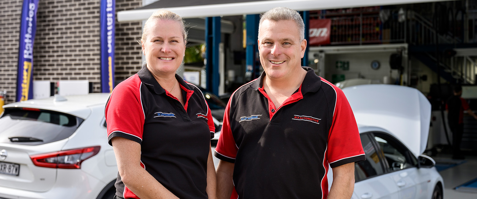 Two automotive technicians, Capricorn Members, in black and red uniforms standing outside a workshop, with two cars and branded signage in the background.