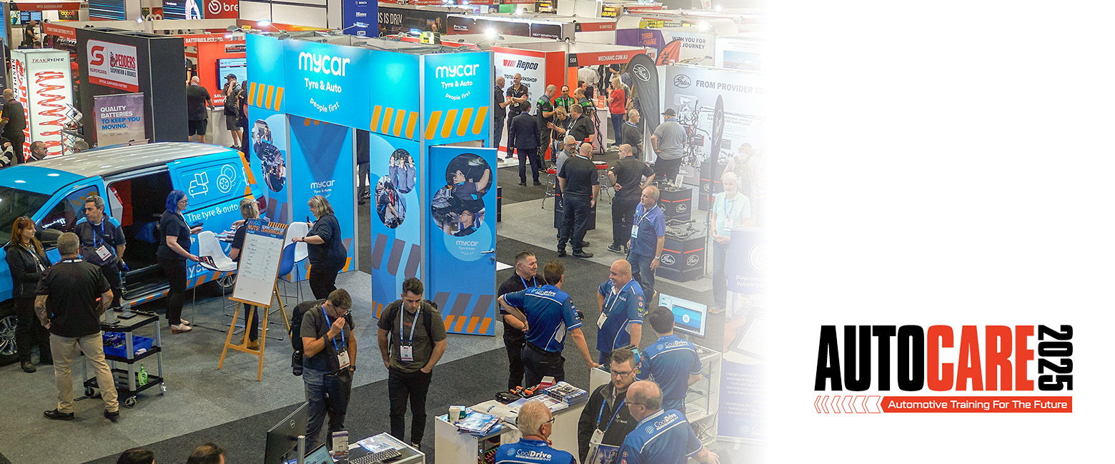 A large crowd of people exploring an auto show (Autocare), with colorful displays and vehicles in the background.