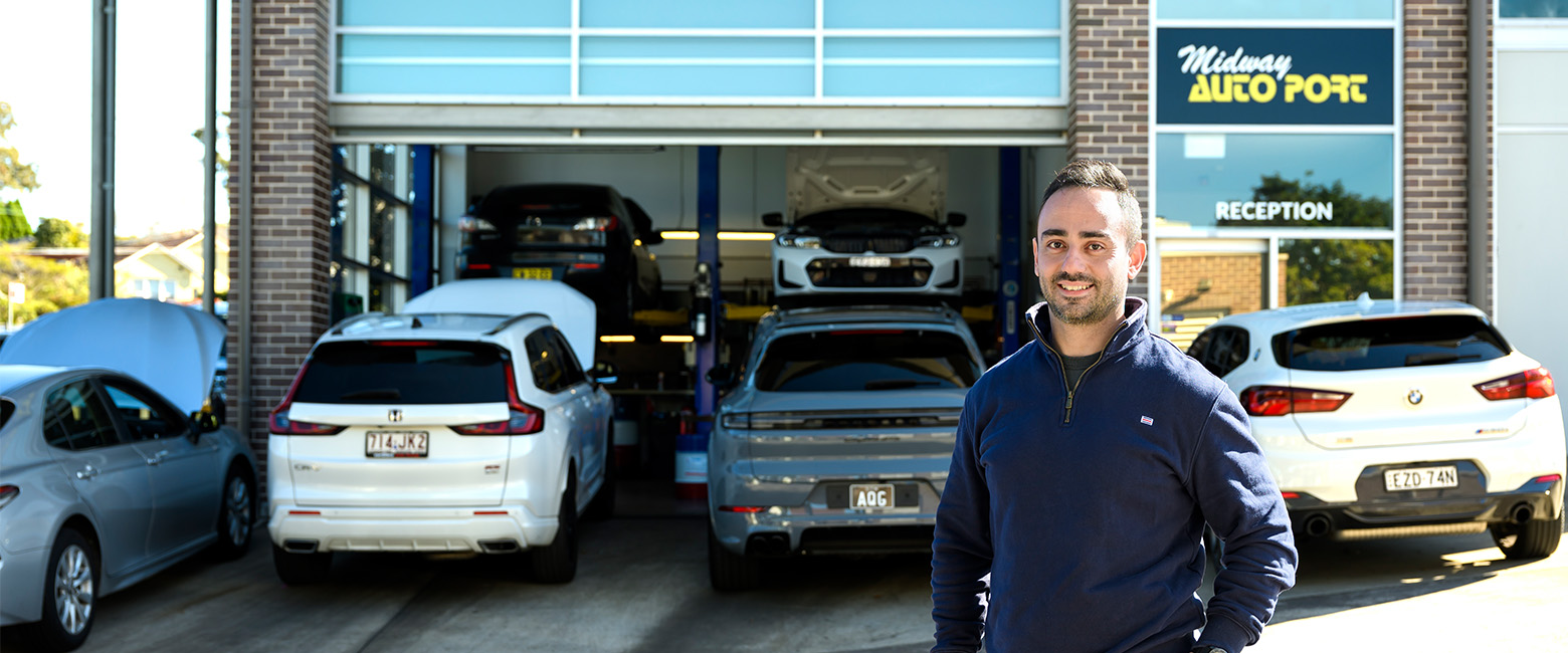 Ross standing outside Midway Autoport workshop in Ryde, with the “Reception” sign visible, cars parked out front and two vehicles on lifts inside the garage.