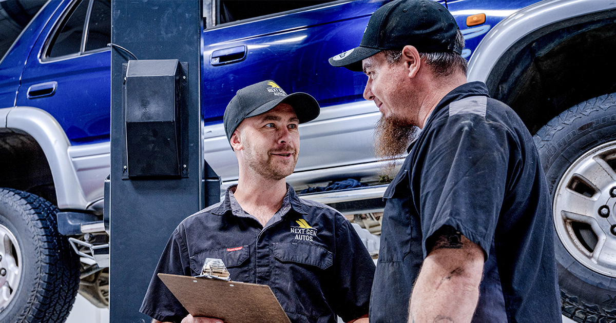 Two automotive technicians in a workshop, engaged in a conversation, with a lifted blue SUV in the background.
