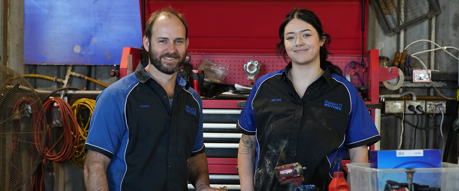 Two automotive technicians in uniforms standing in a workshop, smiling with tools and equipment in the background.