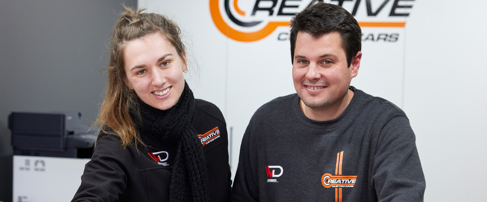 Two team members from a Capricorn Member, Creative Custom Cars, reviewing a document at a front counter, with the company logo visible in the background and office equipment behind them.