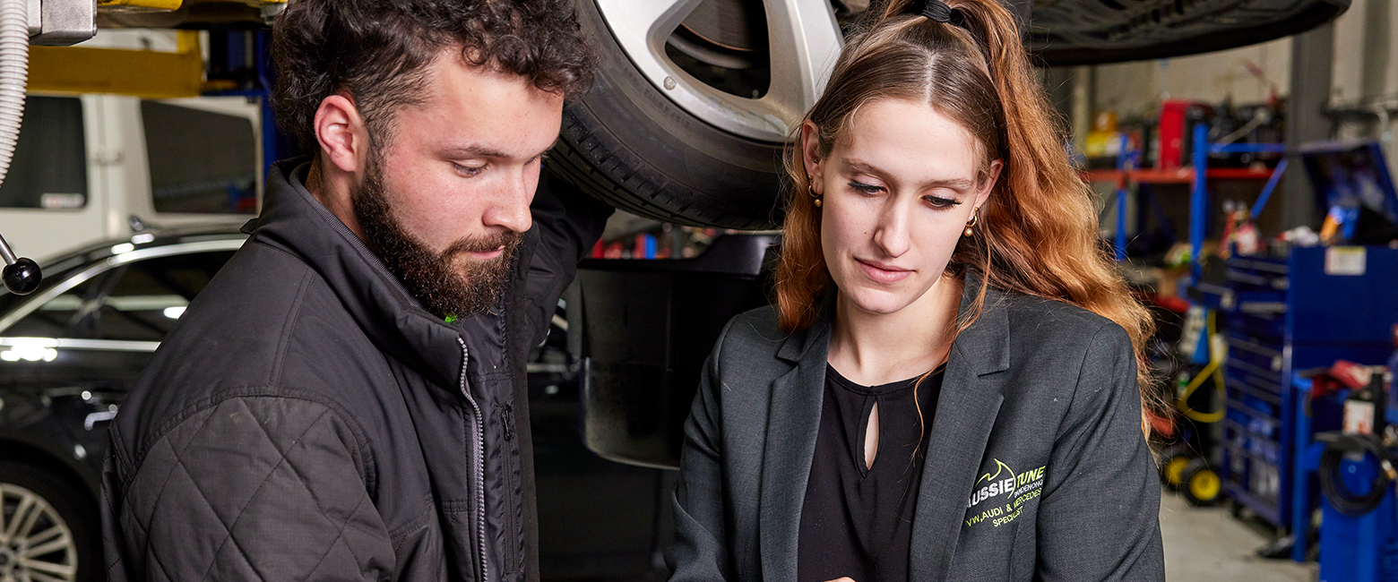 A man and woman in a garage, inspecting a car while examining something on a tablet.