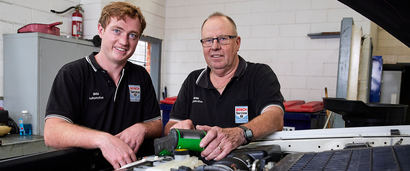 A young apprentice working with an experienced mechanic in a car engine and smiling to the camera