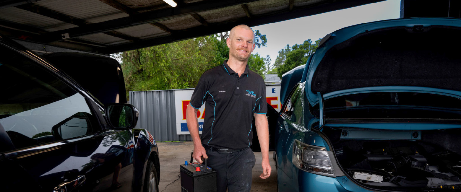 Darren standing in a workshop holding a car battery, wearing a black polo shirt with blue accents. Two cars with open bonnets are visible, and the background shows greenery and a metal-sided building.