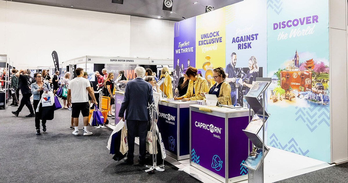 A vibrant exhibition booth with Capricorn branding, featuring team members engaging with Members and Suppliers. The booth displays colorful banners with messages like 'Together We Thrive,' 'Unlock Exclusive Benefits,' and 'Discover the World.'