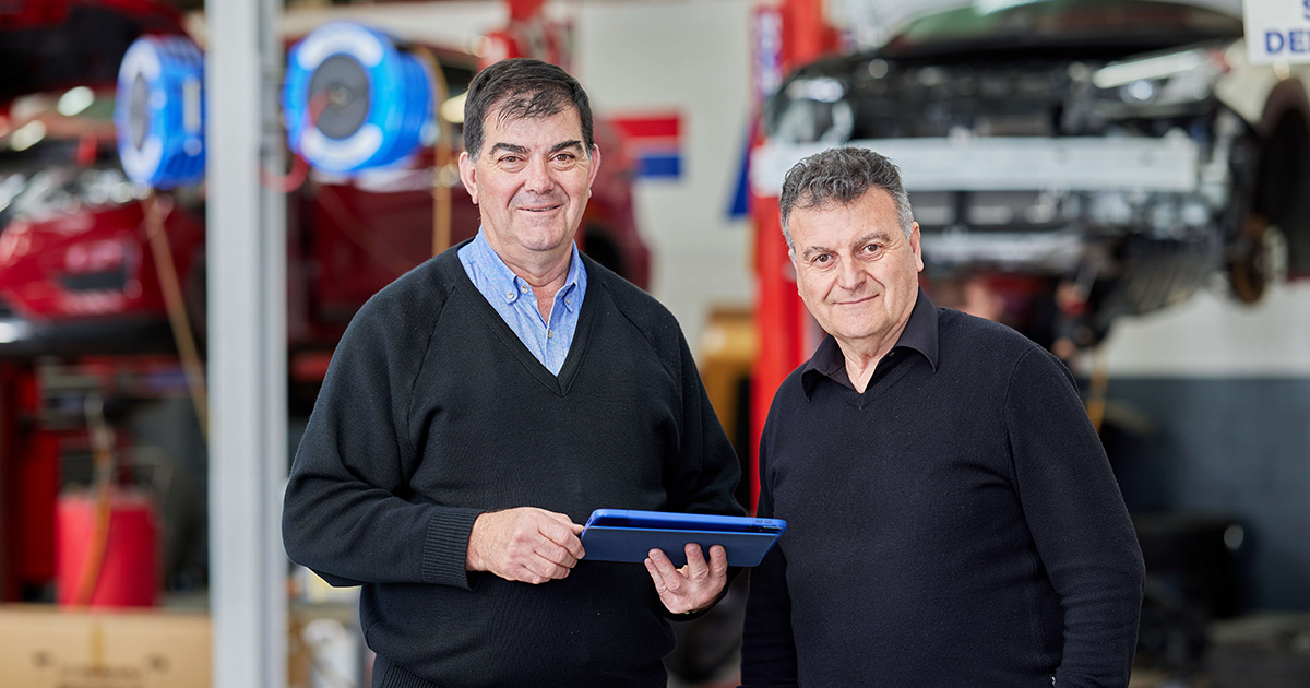 Two Capricorn Members in a vehicle repair shop, engaged in conversation while holding a tablet.