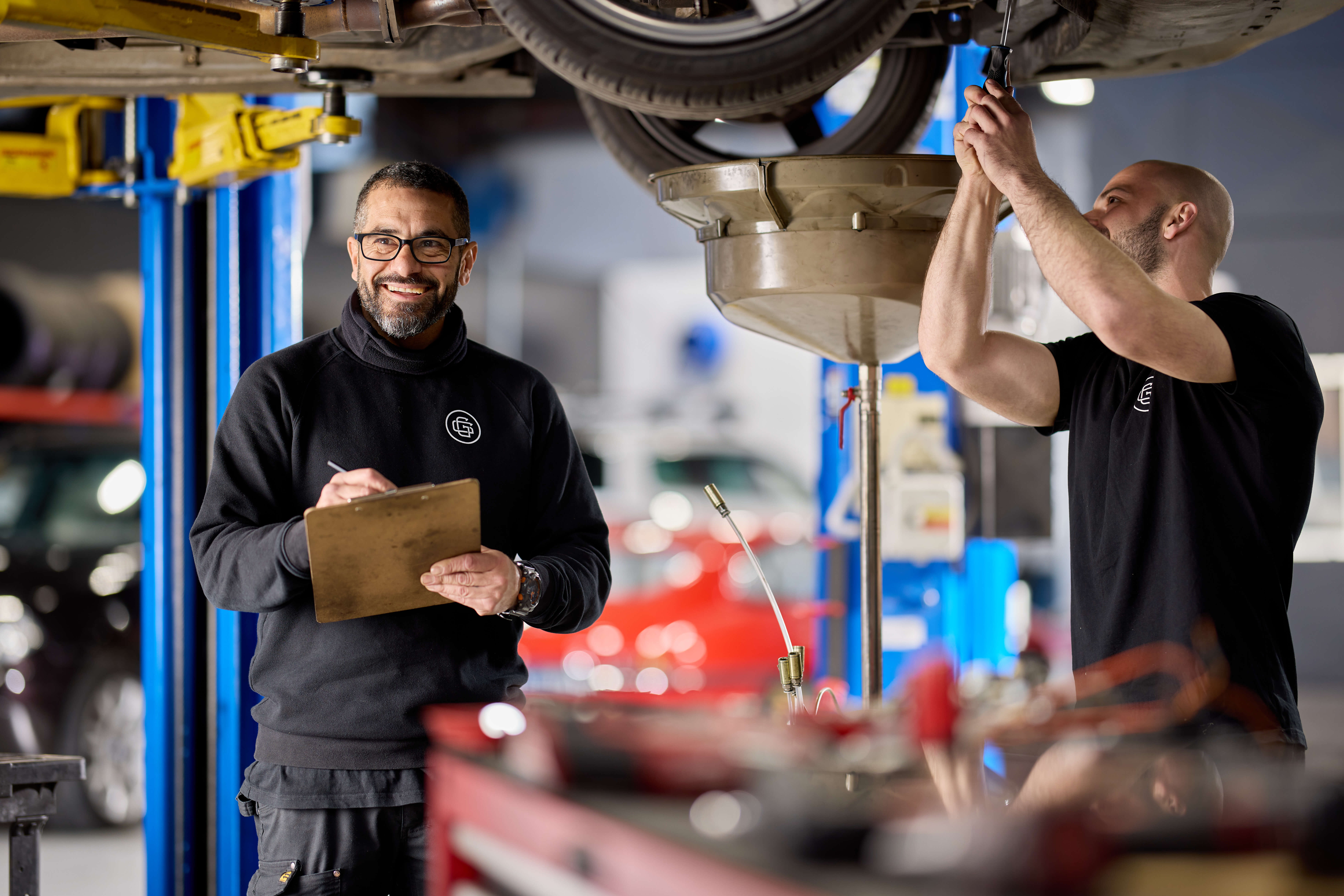 Workshop manager holding clipboard while mechanic works on car beside