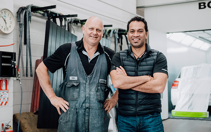 Two auto technicians standing side by side in garage/workshop, both smiling and looking towards the camera